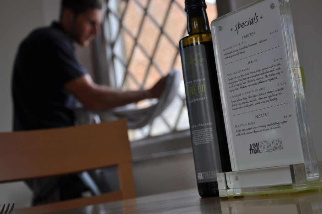 A restaurant table features a bottle of olive oil and a specials menu in focus. In the background, slightly blurred, someone tends to window cleaning with care and precision.