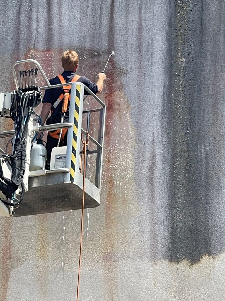 A person in a safety harness expertly navigates a lift platform, using a power washer for render cleaning on the large, weathered wall. Streaks of water and grime map the surface as they work meticulously to restore its appearance.